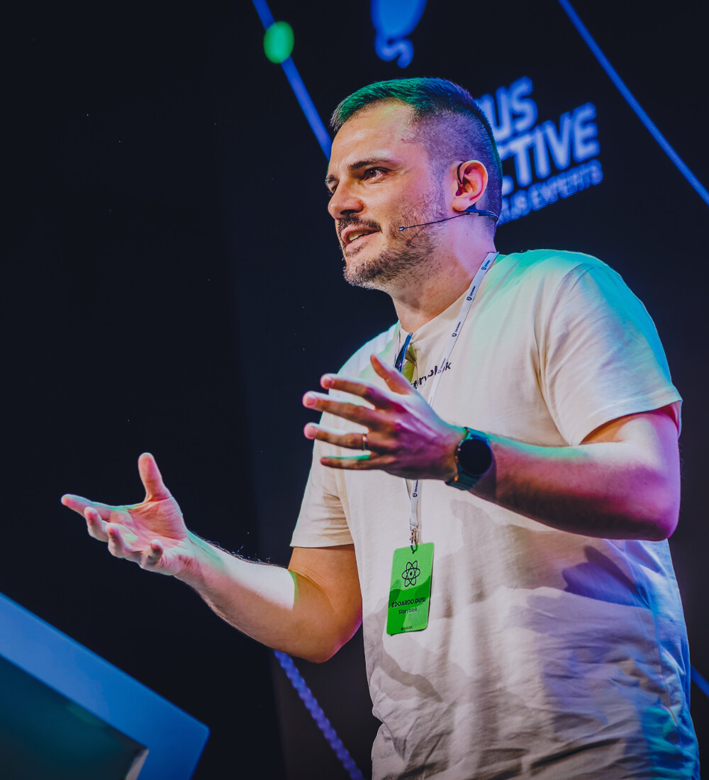 Edoardo Dusi speaking on stage, wearing a headset microphone and mid-gesture.
