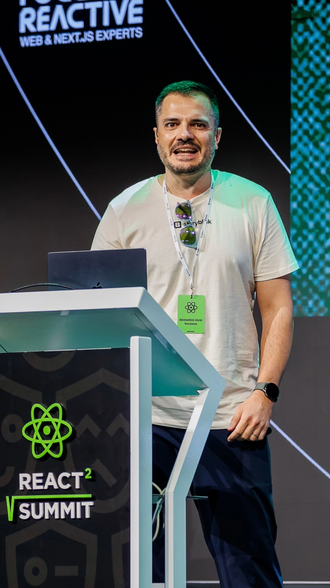 On stage at React Summit, standing beside a branded lectern in front of a large screen.