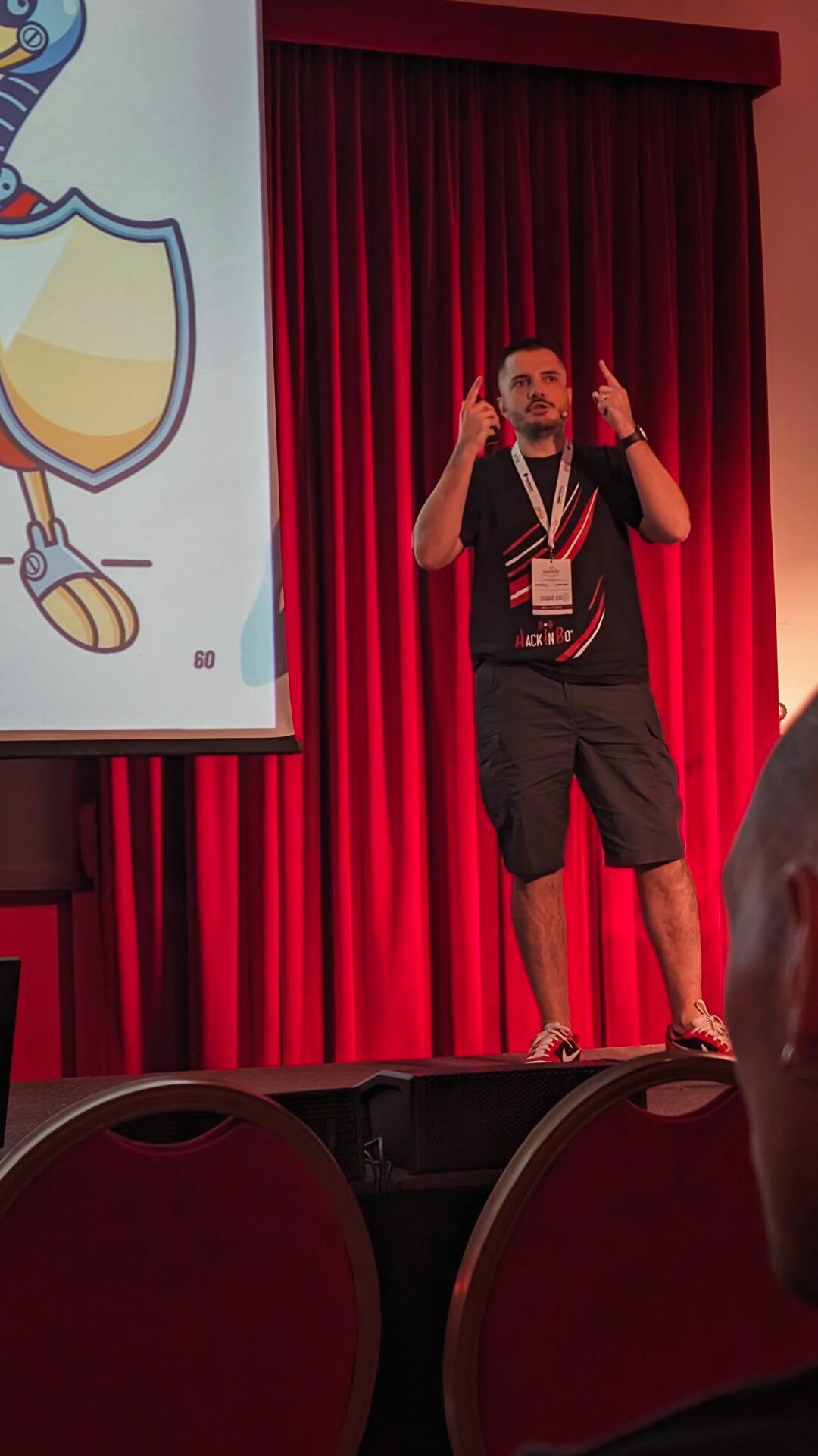 On a red-curtained stage with both hands raised, mid-explanation.