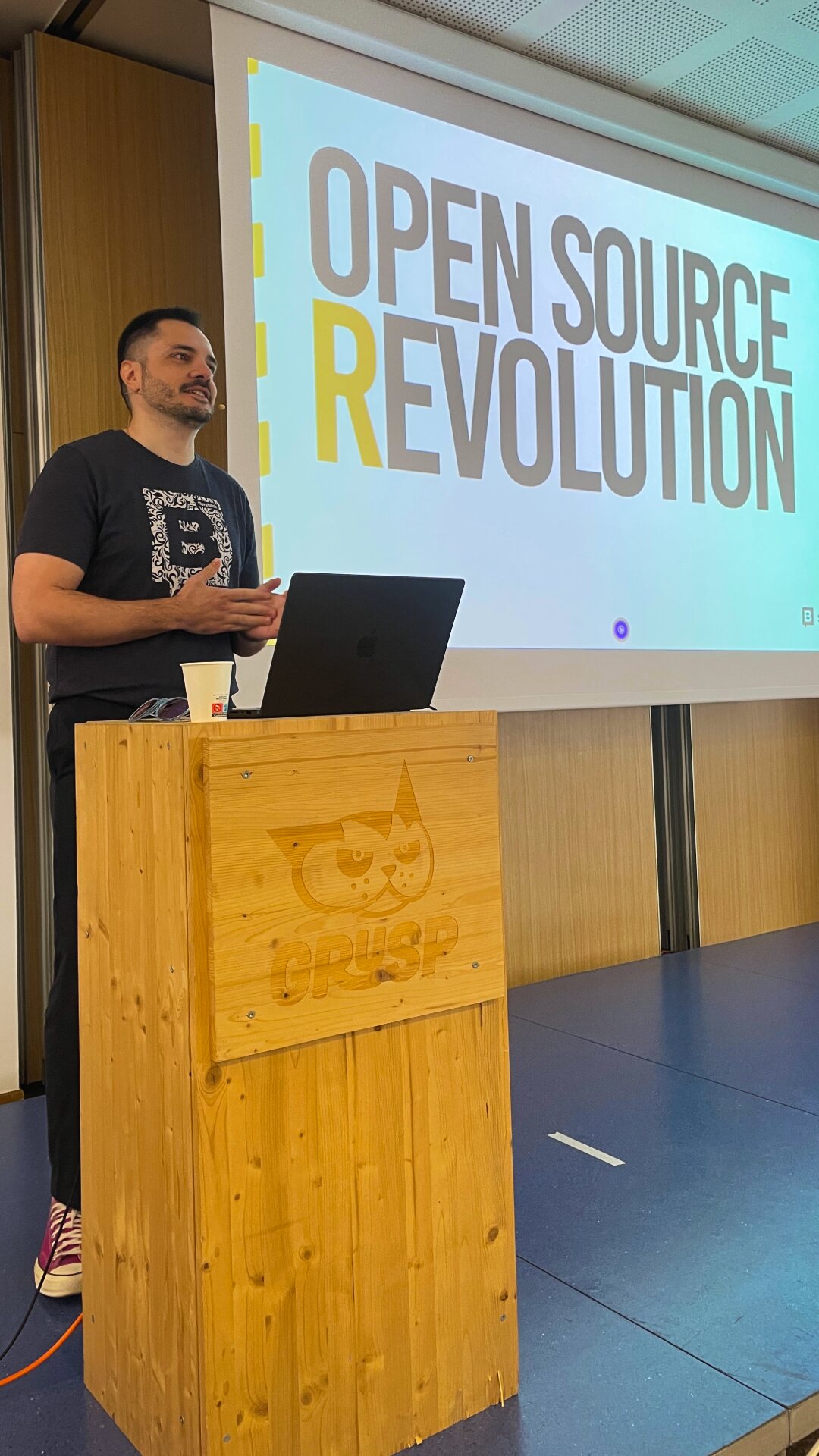 At a wooden lectern beside a slide reading “Open Source Revolution”.
